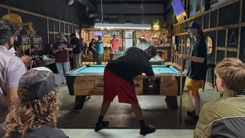 A group of people gathered around a pool table inside a dimly lit bar or lounge, with one person leaning over the table and others watching or standing nearby.